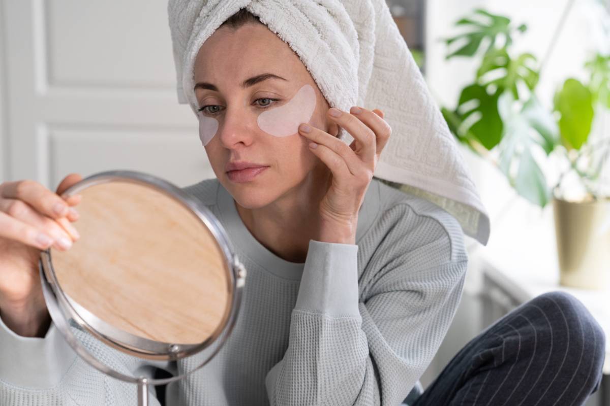 Woman with towel on head apply hydrogel under-eye recovery patches at home, looking in mirror.